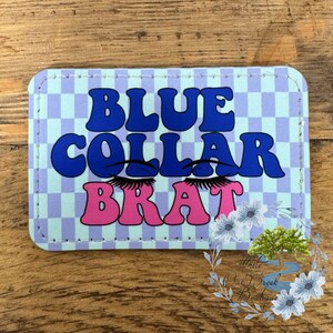 May include: A blue and pink patch with a checkered background. The patch says "BLUE COLLAR BRAT" in a retro font with eyelashes above the word "COLLAR".
