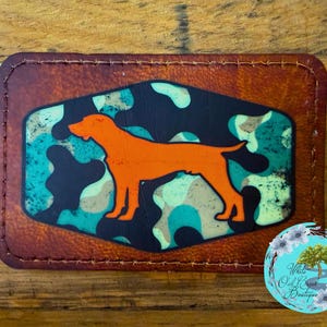 May include: A rectangular patch with a brown leather border. The center features a camouflage pattern in shades of blue, green, and tan, with an orange dog silhouette. The patch is stitched around the edges.