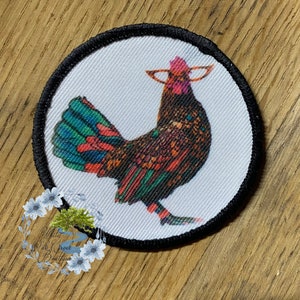 May include: A colorful rooster with a red comb and wattle, green and blue feathers, and a black eye, is printed on a white round patch with a black border.