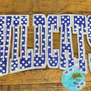May include: A large decorative patch spelling the word WILDCATS in block letters. The letters feature a blue and white patterned sequin design with a silver glitter border. A circular logo with a tree graphic is positioned at the bottom right.