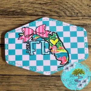 May include: A turquoise and white checkered background with a pink bow and a cartoon outline of the state of Florida. The outline is filled with a colorful beach scene and the text "Florida FL" is written in white.