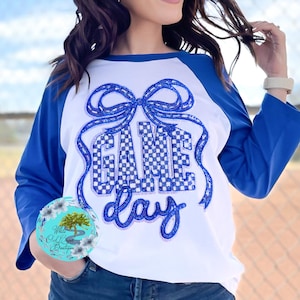 May include: White and blue raglan-style shirt with the words "GAME DAY" in a blue checkered pattern. The shirt has blue sleeves and a large bow design above the text. The shirt is a casual style.
