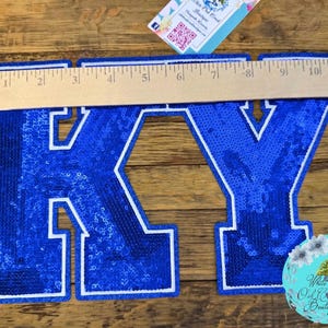 May include: A blue sequined "KY" applique, approximately 11 inches wide, with a white border. The letters are filled with shiny blue sequins. A wooden ruler is placed across the letters. A small, round, floral-themed logo is in the lower right corner.