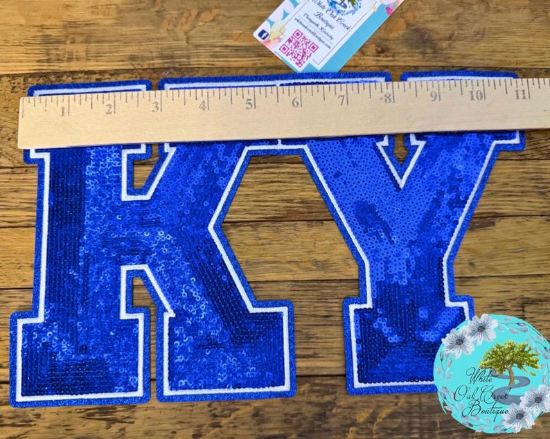 May include: A blue sequined "KY" applique, approximately 11 inches wide, with a white border. The letters are filled with shiny blue sequins. A wooden ruler is placed across the letters. A small, round, floral-themed logo is in the lower right corner.
