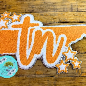 May include: Orange and white Tennessee state-shaped patch with the letters "TN" in white. The patch has a silver glitter border and several glittery white stars. A round blue logo with text is in the lower left corner.