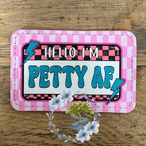 May include: A pink and black checkered background with a white border and a white rectangle in the center. The rectangle has a blue lightning bolt on the left and right sides. The text "HELLO I'M PETTY AF" is written in blue letters inside the rectangle.