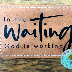 May include: A rectangular, tan-colored patch with stitched edges. The patch features the words "In the Waiting God is working" in black script. A floral logo is in the bottom right corner.