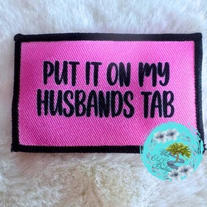 May include: Pink fabric patch with black stitching and the text "PUT IT ON MY HUSBANDS TAB" in black lettering.