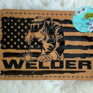 May include: A rectangular leather patch featuring a stylized illustration of a welder in front of an American flag. The word "WELDER" is prominently displayed. The patch is tan with black accents and stitching.