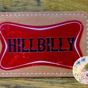 May include: A brown leather patch with white stitching around the edges. The patch has a red, white, and black design with the word "HILLBILLY" in black letters.