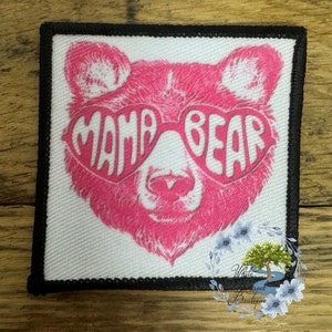 May include: Square patch with a pink bear wearing heart-shaped sunglasses. The sunglasses spell out "MAMA BEAR" in white letters. The patch has a black border and is on a white background.