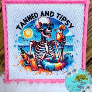 May include: A white square patch with pink stitching around the edges. The patch features a cartoon skeleton wearing sunglasses and holding a tropical drink. The text "Tanned and Tipsy" is written in a black, cursive font around the skeleton.
