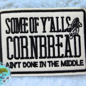 May include: A rectangular embroidered patch with a cream-colored background and a thick black border. The black embroidered text reads SOME OF Y'ALLS CORNBREAD AINT DONE IN THE MIDDLE, accompanied by a small black embroidered corn cob icon.