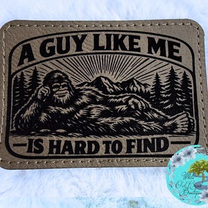 May include: A rectangular, tan-colored patch with the text "A GUY LIKE ME IS HARD TO FIND" in black. The patch features a black illustration of a Bigfoot-like creature relaxing in front of mountains and trees.