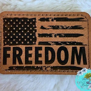 May include: A rectangular, brown leather patch featuring a distressed American flag design. The flag's stars and stripes are visible, with the word "FREEDOM" boldly printed beneath. The patch has a stitched border.