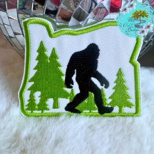 May include: Green and white embroidered patch shaped like the state of Oregon with a black silhouette of Bigfoot walking through a forest.