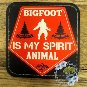 May include: A black and red warning sign with white text that reads "BIGFOOT IS MY SPIRIT ANIMAL". The sign features a silhouette of Bigfoot standing in a forest setting.