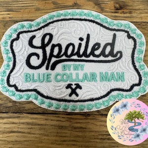 May include: White embroidered patch with a teal border. The patch features the text "Spoiled by my Blue Collar Man" with a pair of crossed hammers underneath.