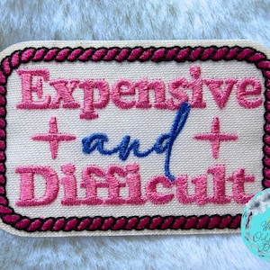 May include: A rectangular embroidered patch with a cream-colored background and a dark pink rope-style border. The text reads Expensive in pink, and in blue, and Difficult in pink, with two small pink cross symbols flanking the word and.