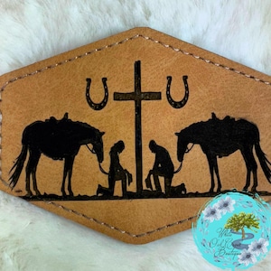 May include: A tan leather patch with a stitched border. The patch features a black silhouette design of two horses, two kneeling figures, a cross, and two horseshoes. The words "White Oak Creek Boutique" are in a blue circle.