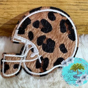 May include: A brown and black leopard print football helmet patch with white stitching. The patch is on a white fluffy surface and a wooden background. The patch is a decorative item.