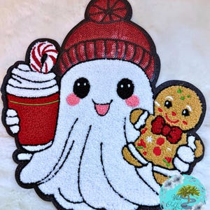 May include: A white ghost-shaped plushie wearing a red sequined beanie, holding a red cup of whipped cream and a gingerbread man. The ghost has rosy cheeks and a smiling face. The plushie has a black outline.