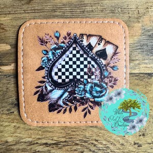 May include: A brown leather coaster with a checkered ace of spades design, surrounded by blue roses and feathers. The coaster has a stitched edge.