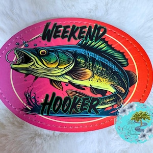 May include: Oval-shaped patch with a vibrant design featuring a detailed illustration of a fish with an open mouth, a fishing hook, and the words "WEEKEND HOOKER" in bold letters. The patch has a pink and red color scheme.