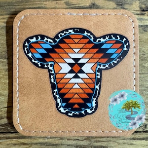 May include: A brown leather coaster with a geometric design of a cow head in orange, white, and blue colors. The coaster has a white stitched border.