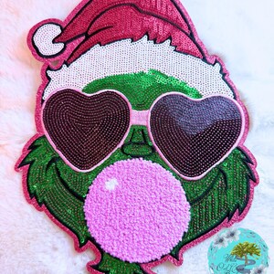 May include: A whimsical Christmas-themed applique featuring a green character with heart-shaped sunglasses and a pink bubblegum bubble. The applique is embellished with sequins in shades of green, pink, and brown, and a Santa hat.