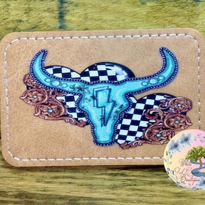 May include: A light blue cow skull with a lightning bolt design is surrounded by a black and white checkered pattern and brown floral accents. The design is on a brown leather patch.