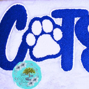 May include: A tufted wall hanging featuring the word CATS in deep blue, with a white paw print replacing the letter A. The letters have a thick white border and a textured, plush surface. A circular logo with floral art and text is visible.