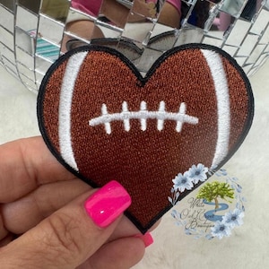May include: A heart-shaped patch featuring a football design. The patch is brown with white stitching and a black border. The patch is held in front of a mirrored disco ball.