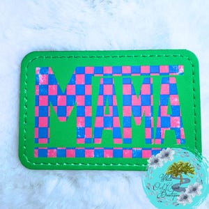 May include: A rectangular patch with the word "MAMA" in a pink and blue checkered pattern on a green background. The patch has a stitched border and a logo in the bottom right corner.