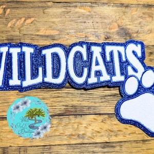 May include: A blue and white Wildcats design with a paw print. The word "WILDCATS" is in white block letters with a blue glitter outline. A blue paw print is next to the word. A small round logo is in the lower left corner.