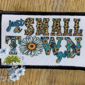 May include: A white patch with black stitching around the edges. The patch features a colorful design with the text "Just a Small Town Girl" in a variety of fonts and colors.