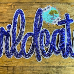 May include: A blue and silver Wildcats sign with a cursive font. The letters are filled with blue sequins and outlined in silver. The sign is on a wooden background.
