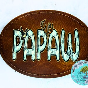 May include: A brown, oval-shaped patch with the word "PAPA" in large, light blue camouflage letters. The letter "W" has a hook design. A bird and antlers are incorporated into the design. The patch has a stitched border.