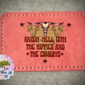 May include: Pink leather patch with a brown stitched border. The patch features a gold silhouette of a skull with horns and cacti. The text "Raisin' Hell With The Hippies And The Cowboys" is printed in brown.