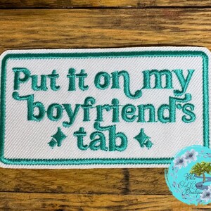 May include: White rectangular patch with teal embroidered text that reads "Put it on my boyfriend's tab". The patch has a teal border and two teal diamond shapes. The patch is on a wooden surface.