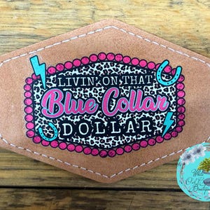 May include: A brown leather patch with the text "LIVIN ON THAT Blue Collar DOLLAR" in pink and white, surrounded by a pink beaded border and turquoise accents. The patch has a stitched border and a leopard print background.
