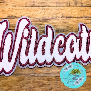 May include: A white and maroon "Wildcats" patch with a gray border, displayed on a wooden surface. The word "Wildcats" is in a cursive font. A teal circular logo with white flowers and text is in the bottom right corner.