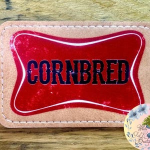May include: A brown leather patch with a red and white border and the word "CORNBRED" in black lettering.