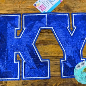 May include: Blue sequined patches spelling "KY" with white borders. The patches are on a wooden surface. The image also includes a business card and a logo for "White Oak Creek Boutique".