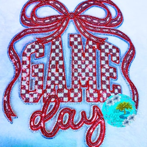 May include: A decorative "Game Day" sign with red sequins and a checkered pattern. The sign features a large bow at the top and the words "Game Day" in a playful font. A small, round logo is visible in the lower right corner.