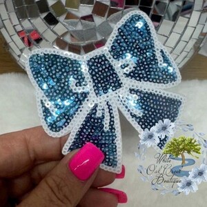 May include: A large blue and white sequined bow with a white outline. The bow is made of sequins and has a shiny finish.