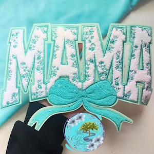 May include: A white and teal "MAMA" patch with floral detailing and a teal sequined bow. The patch has a teal border and is set against a teal background. The White Oak Creek Boutique logo is in the lower right corner.