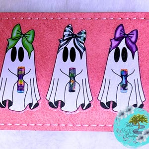 May include: Pink rectangular patch featuring three cartoon ghosts with black eyes and holding colorful objects. Each ghost wears a different colored bow: green, black and white striped, and purple. The patch has a stitched border.