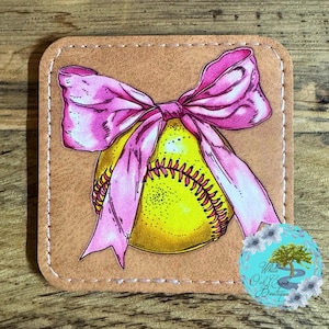 May include: A softball with a pink bow on top. The softball is yellow with white stitching. The bow is made of pink ribbon. The background is a brown leather coaster.
