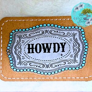 May include: A rectangular, tan leather patch with the word "HOWDY" in bold black letters. The text is inside a decorative, ornate frame with a light blue beaded border. The patch has a stitched border.